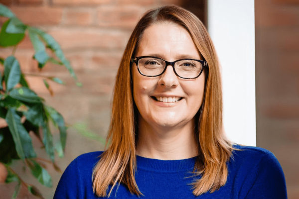A portrait of a smiling Donna Castle, a white woman with shoulder length chestnut hair, wearing glasses and a blue jumper. There is a plant and brick wall in the background