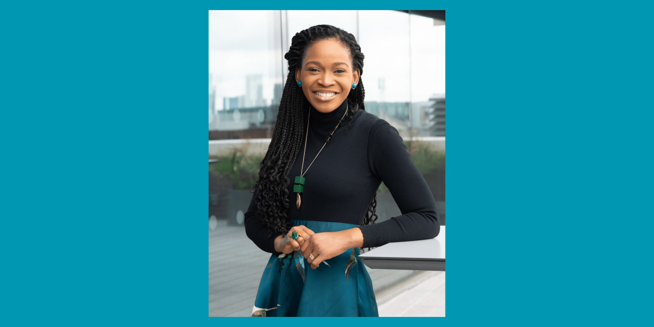 A portrait of a smiling Eduvie Martin, a black woman with long dark hair, dark top and blue skirt, stood on a balcony with buildings behind her