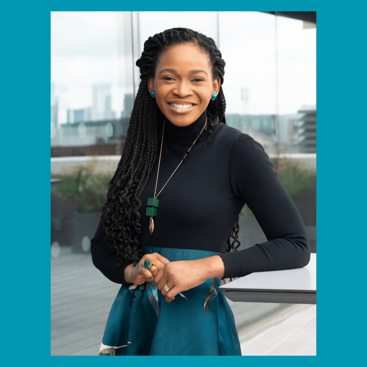 A portrait of a smiling Eduvie Martin, a black woman with long dark hair, dark top and blue skirt, stood on a balcony with buildings behind her