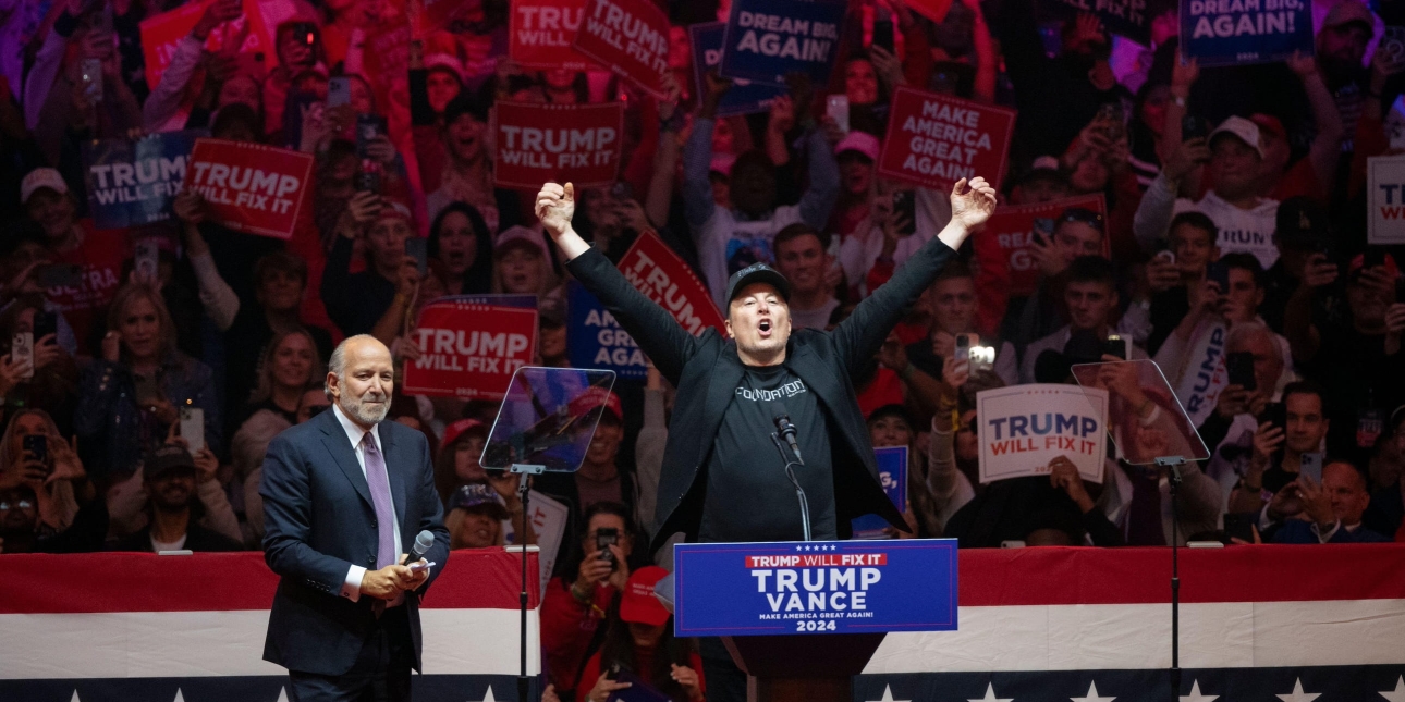 Elon Musk holds his arm up wide, while on stage with another man at a political rally. In the background are supporters holding Trump placards