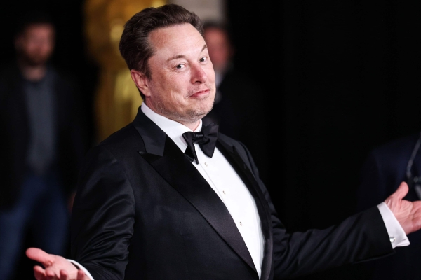 Elon Musk, a white man with short brown hair, looks at the camera with his arms open, while wearing a black suit and bow tie. The background is mostly dark with glimpsed of people behind him