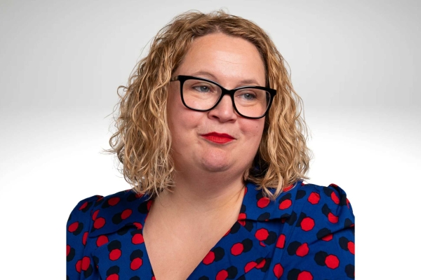 A side portrait of Emma Duke, a white woman with blonde shoulder length hair, who wears glasses and a red and black polka dot blue top