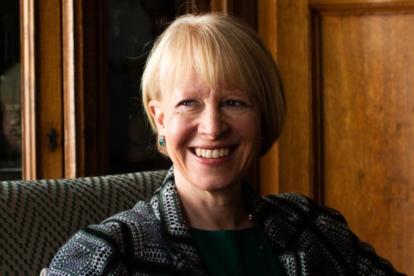 A colour portrait of a smiling Emma Fox. Emma is a white woman with blond hair wearing a green patterned jacket. She is sat on a sofa in front of a display case of wine and a wood panel.