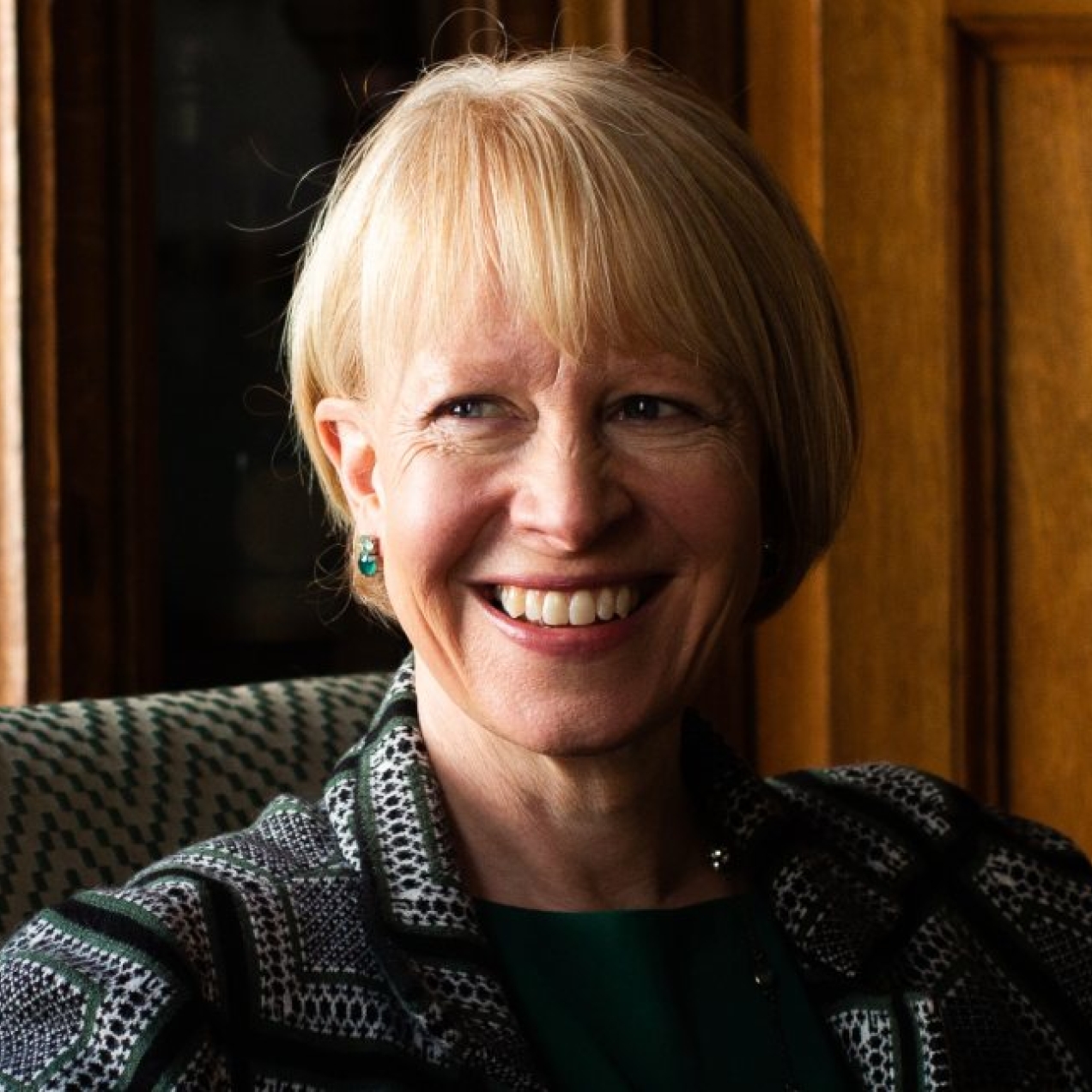 A colour portrait of a smiling Emma Fox. Emma is a white woman with blond hair wearing a green patterned jacket. She is sat on a sofa in front of a display case of wine and a wood panel.