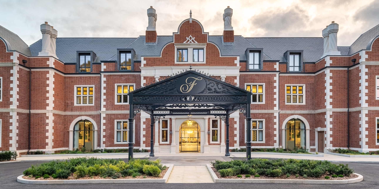 The exterior entrance of Fairmont Windsor Park, an ornate three storey red brick building with a dozen windows, eight chimneys and a decorate black canopy embossed with an italic F covering a portion of the green vehicle turning circle out front. It