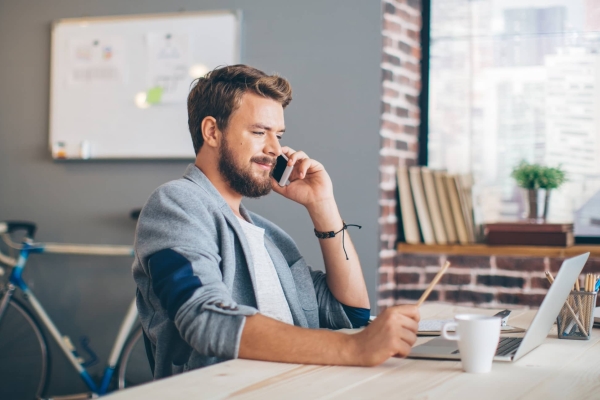 A white man with blond hair, wearing a blue shirt, sits at a desk in a spacious apartment, chatting on his mobile while looking at his laptop. Behind him are a blue bicycle resting against a wall and large window with a city skyline