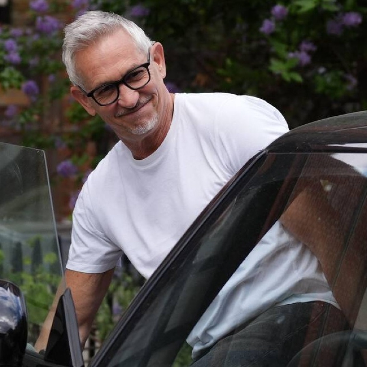 Gary Lineker, dressed in a white t-shirt, getting into the driver's seat of his black car. Lineker is a white man with grey hair and glasses. There are red brick houses in the background.