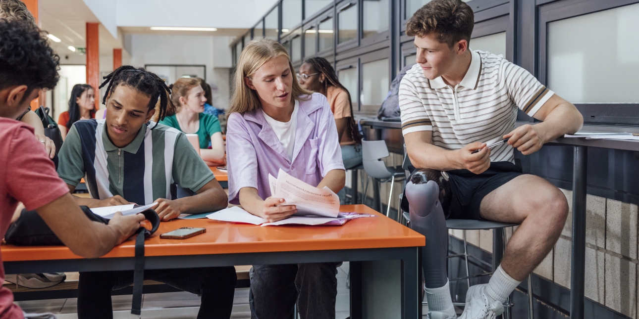 Four college students gathered round a desk. Facing the photo from left to right: a Black male with short hair and green striped shirt reading; a white male with shoulder length blond hair and pink shirt points to paperwork; a white male with short b