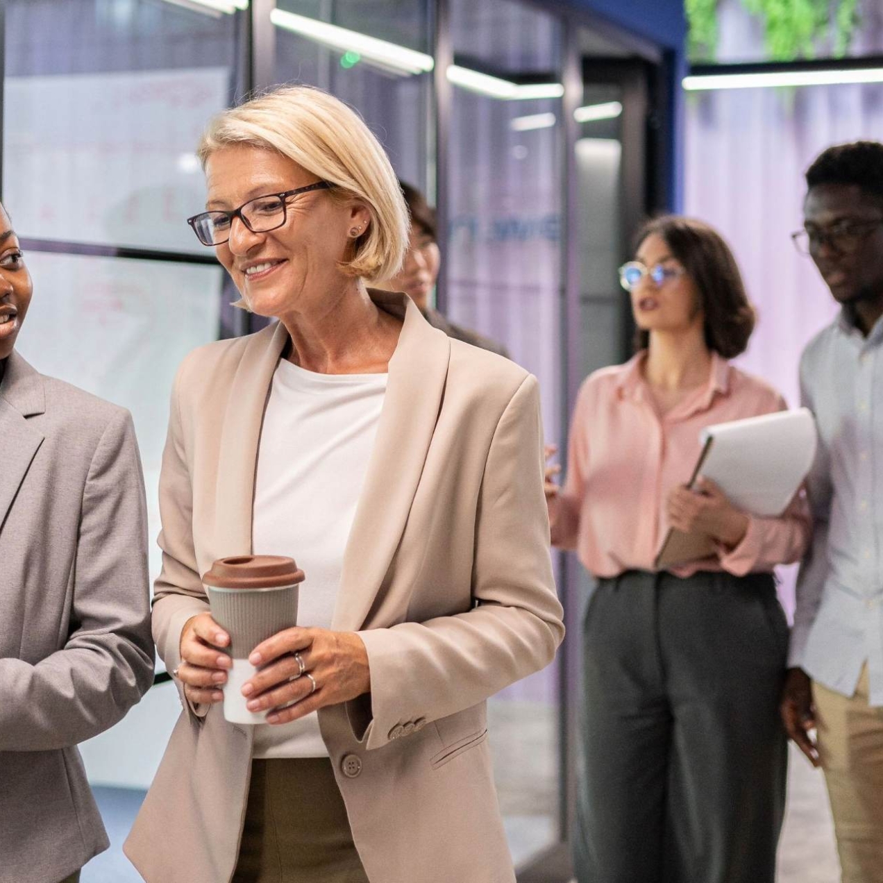 Walking through an office corridor, a young black business woman holding a book in deep conversation with an older white business woman holding a coffee cup.