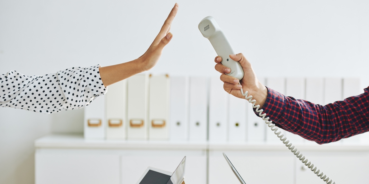 A white hand on the right of the photo tries to pass a phone receiver to a colleague over their laptops. The colleague's light-skin hand is held up suggesting that they are refusing to take the call. The person on the left wears a black spotted white