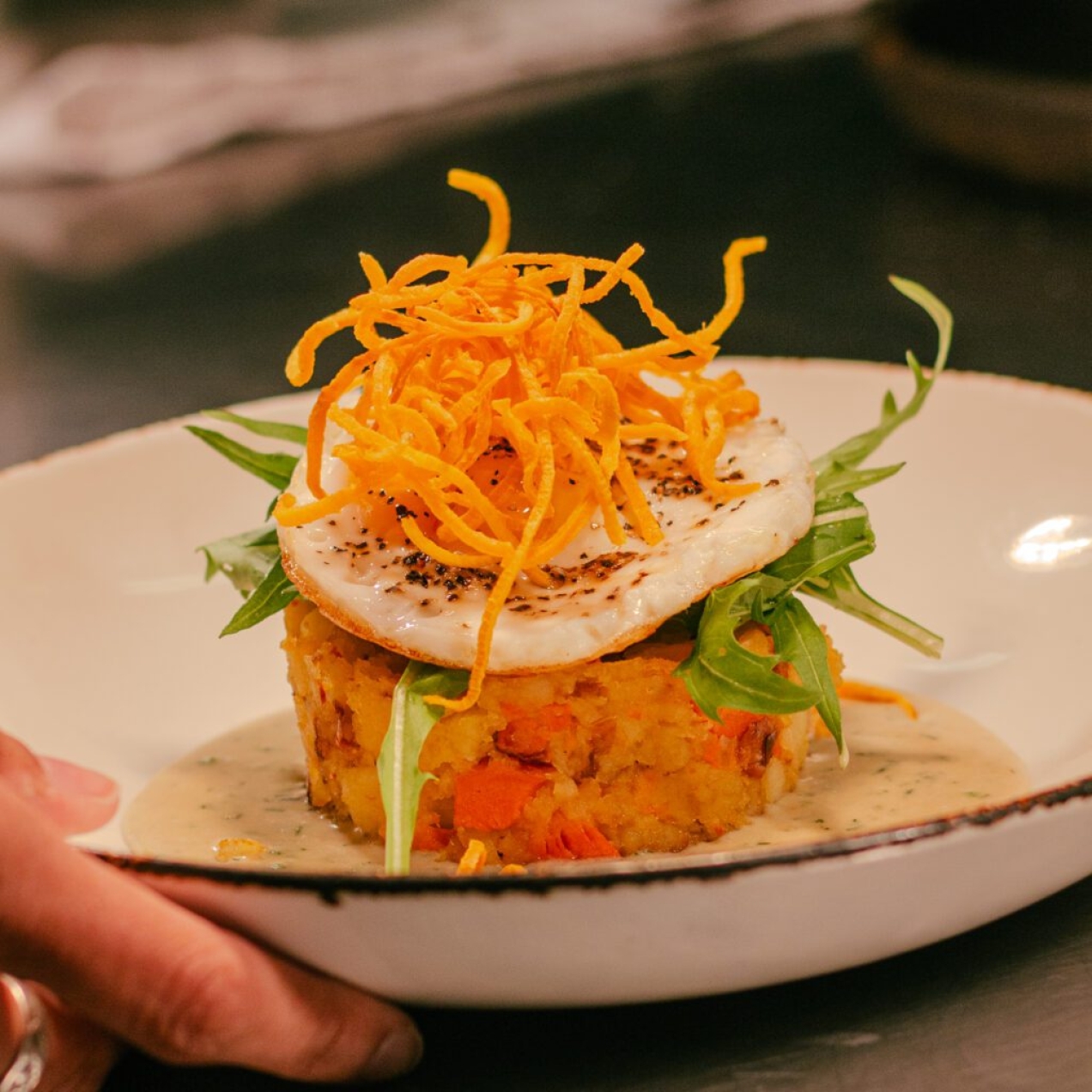 A hand holds a white bowl with Smoked Carrot & Potato Hash, Crispy Hens Egg, Dill Cream