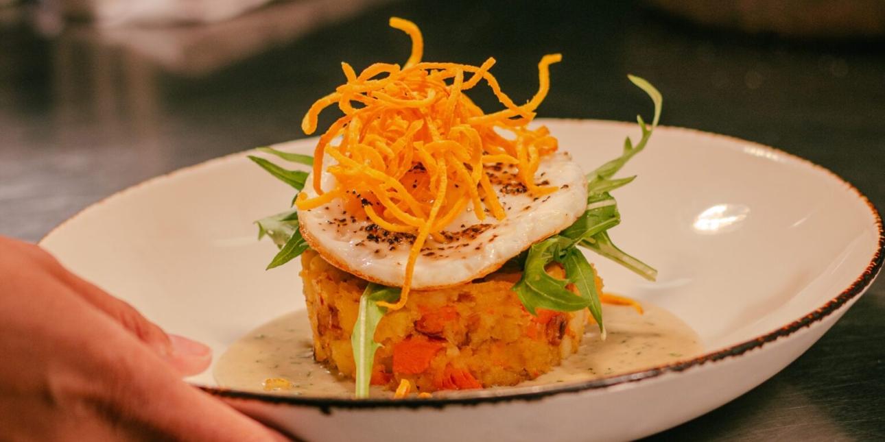 A hand holds a white bowl with Smoked Carrot & Potato Hash, Crispy Hens Egg, Dill Cream