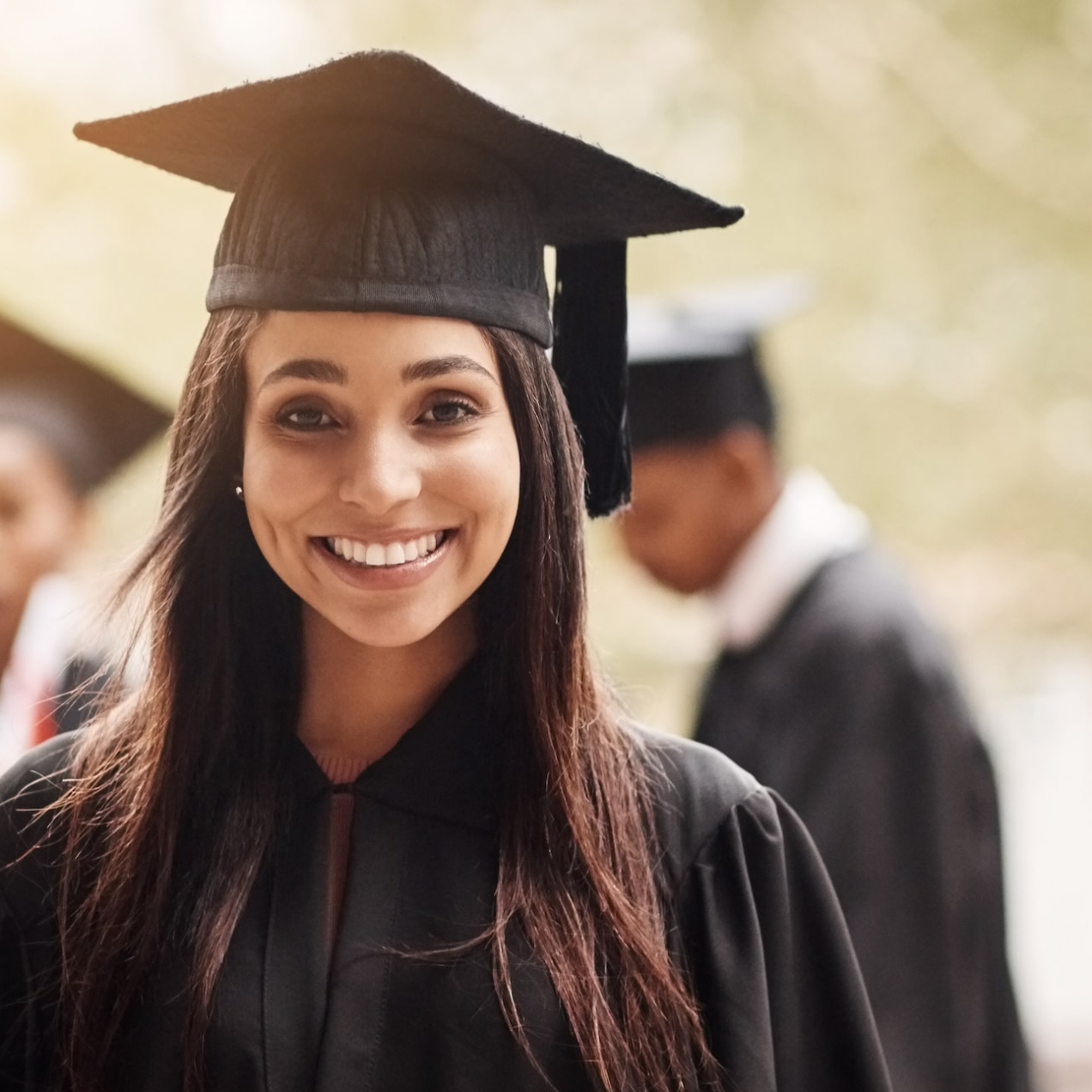 Portrait of a young smiling Asian woman wearing a black graduation gown and mortarboard hat. Out of focus in the background are three fellow graduates