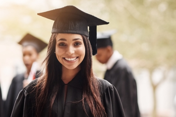 Portrait of a young smiling Asian woman wearing a black graduation gown and mortarboard hat. Out of focus in the background are three fellow graduates