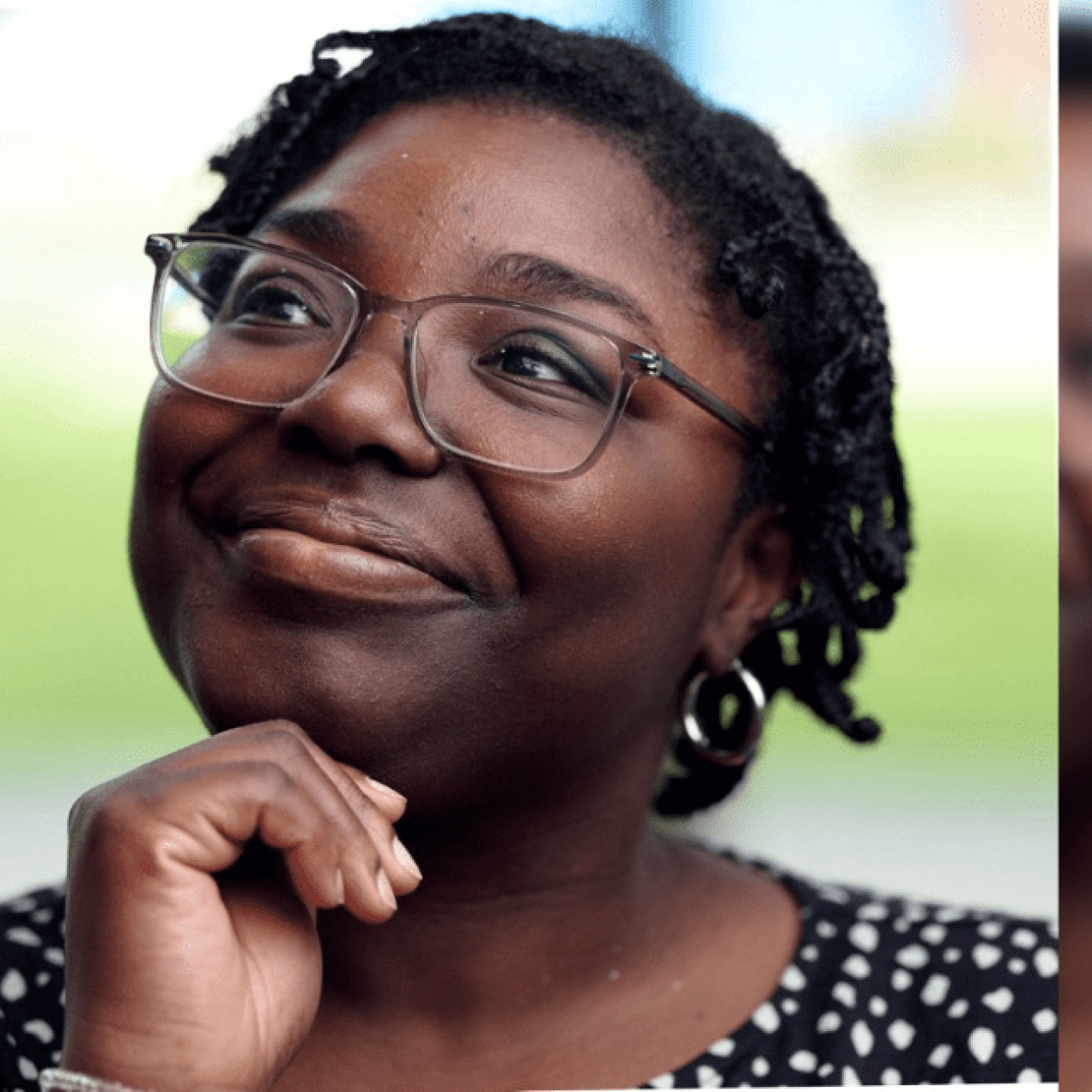 Hannah Abban holding her hand under her chin while looking reflective. Hannah is a Black woman with glasses, short dark hair and wears a white polka dot black top