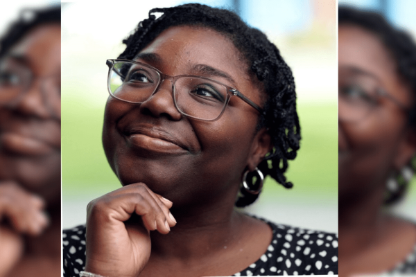 Hannah Abban holding her hand under her chin while looking reflective. Hannah is a Black woman with glasses, short dark hair and wears a white polka dot black top