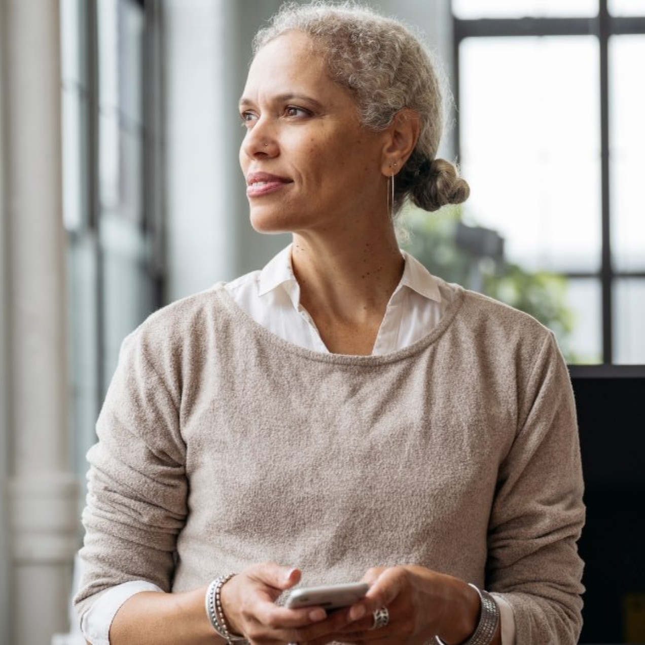 In a light-filled office, a contemplative businesswoman stands with her head faced towards the window while holding a phone in her hand.