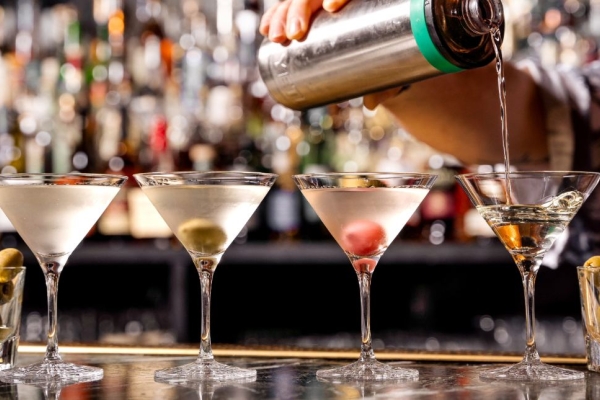 Four glasses of martini on a bar flanked by two small glasses of olives. An unidentified mixologist pour the drink from a silver shaker into the fourth martini glass. The background is out of focus.