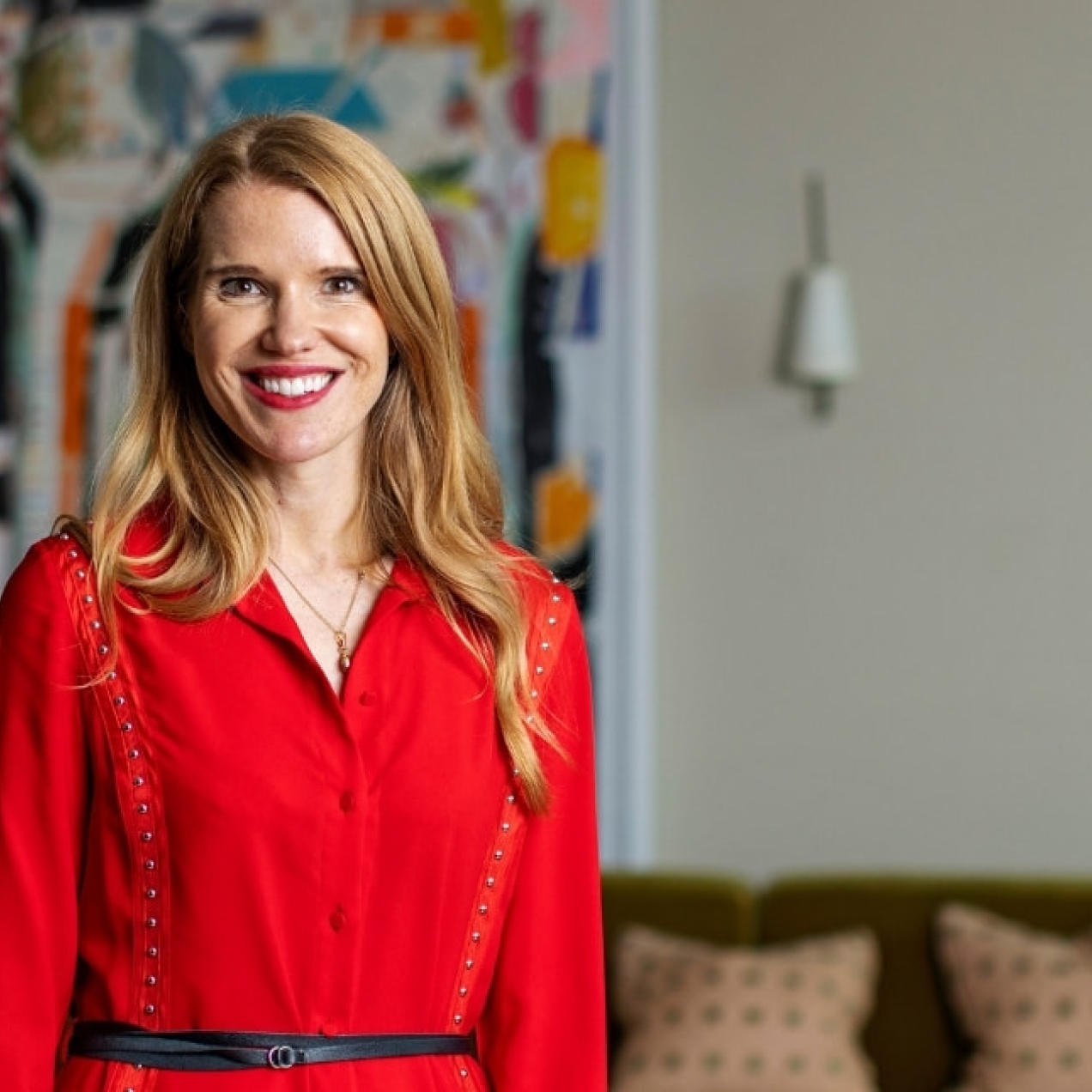 A portrait of Helen Tupper facing the camera in a modern living room while wearing a red dress. Tupper is a white woman with blond hair. Behind her are a wall mounted painting and soft furnishings.