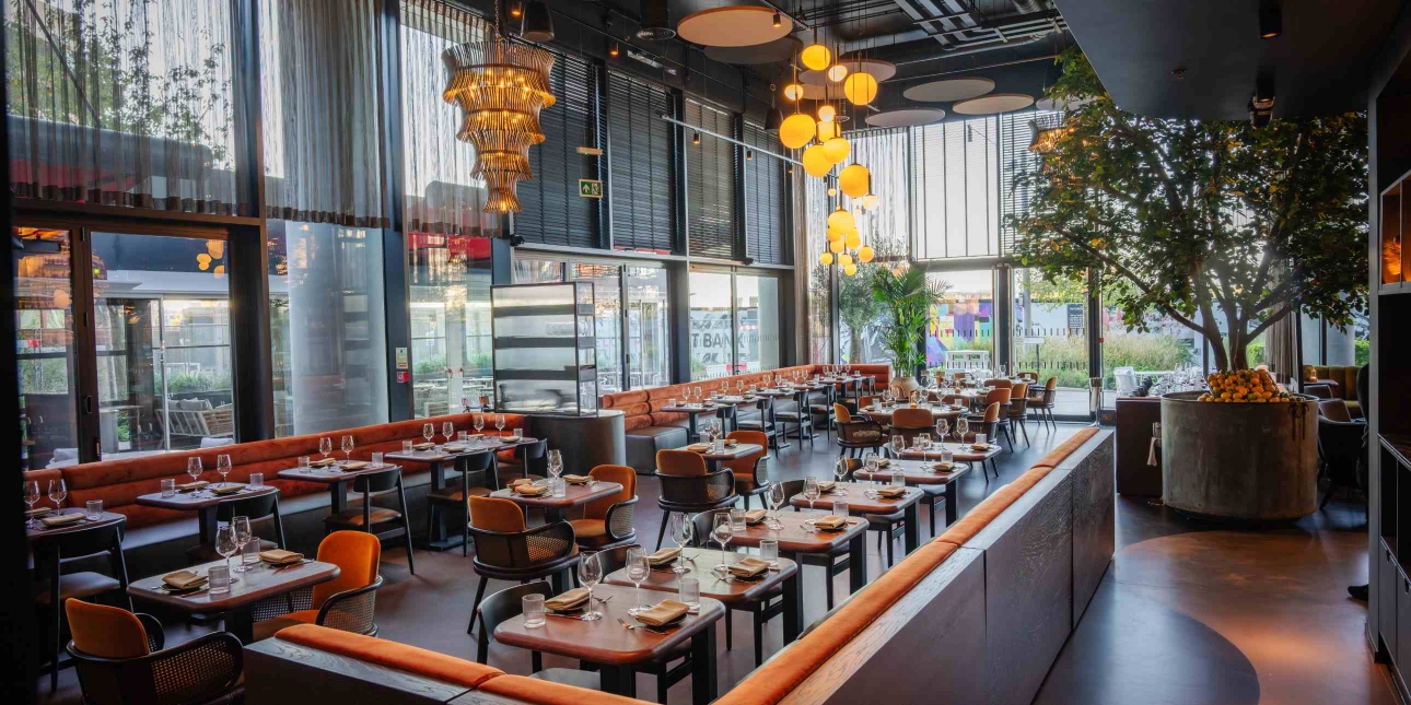 The interior of Hera restaurant with floor to ceiling windows, and a mix of tables, chairs and longitude oranged cushioned seating. There is an olive tree in the top right hand corner and lights hanging from the ceiling.