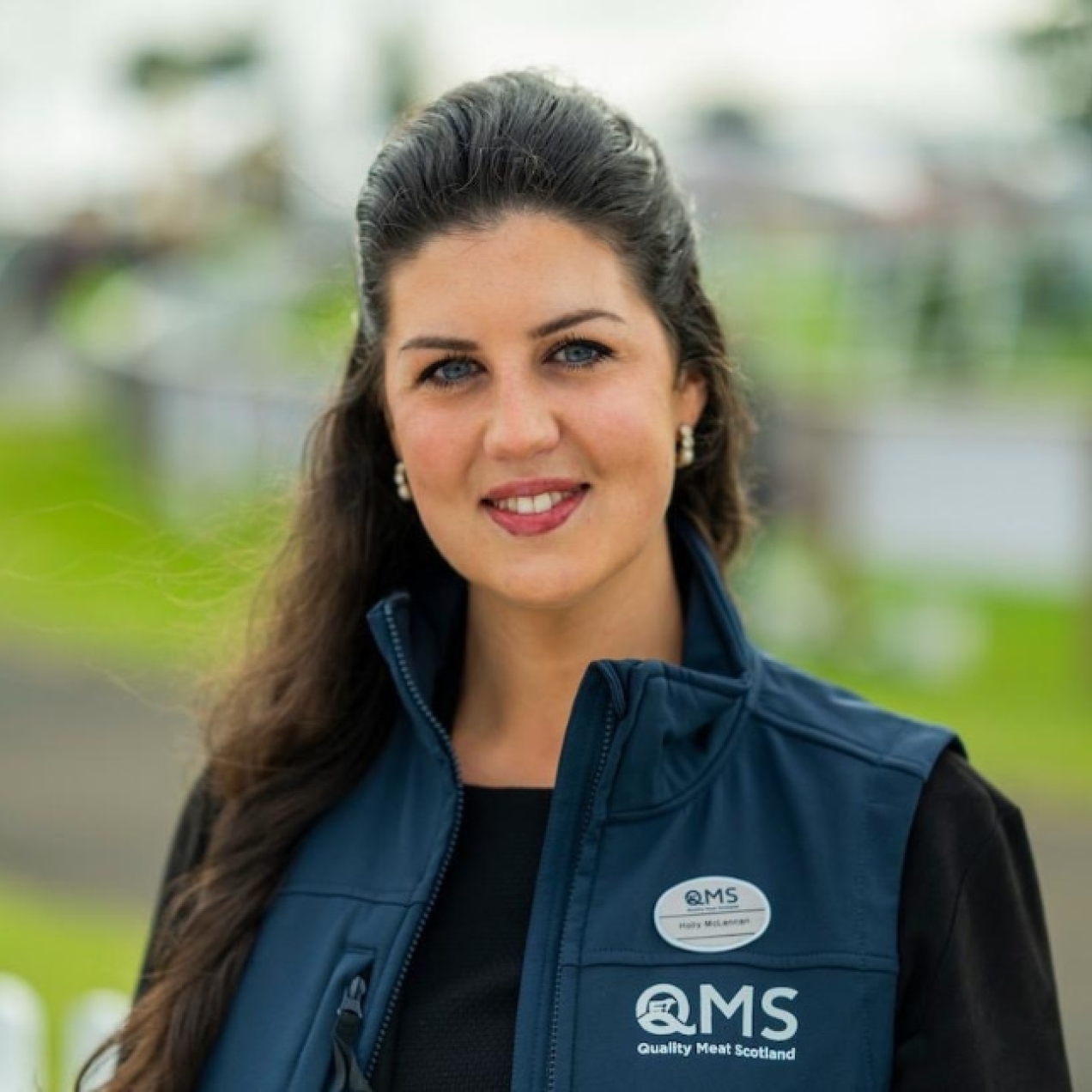 A colour portrait of Holly McLennan. Holly is a white woman with long dark hair wearing a blue gilet with writing on it. There is greenery behind her.