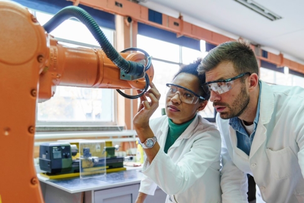 Two colleagues in white lab coats and protective glasses - a Black woman on the left and a white man on the right - inspect an orange robotic arm in a laboratory.