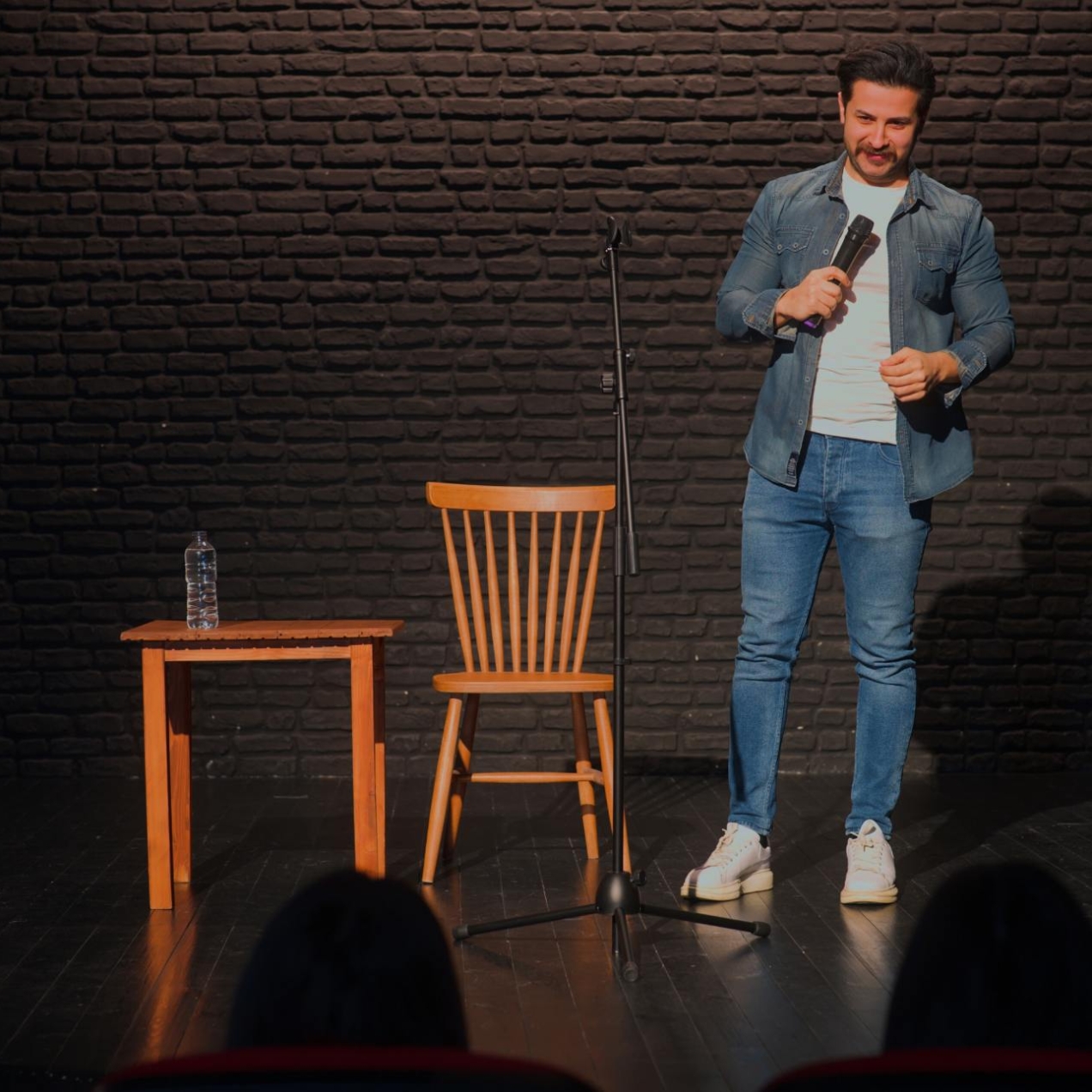 A white man with short dark hair and beard is stood on a stage holding a microphone facing a silhouetted audience