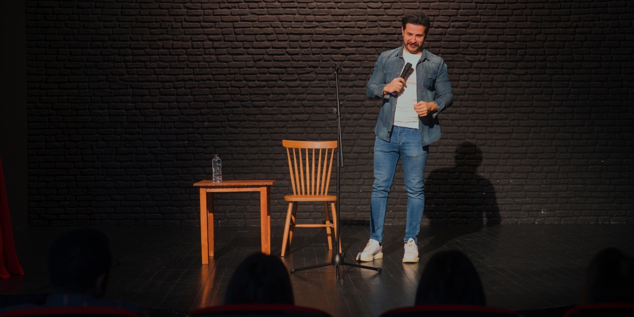 A white man with short dark hair and beard is stood on a stage holding a microphone facing a silhouetted audience