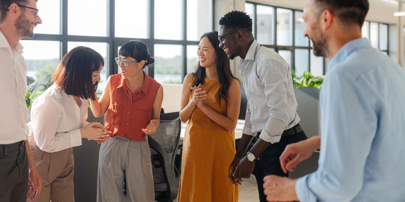 A group of six colleagues stood laughing and joking in front of a large window. Among them are two white men, one Black man and three Asian women.