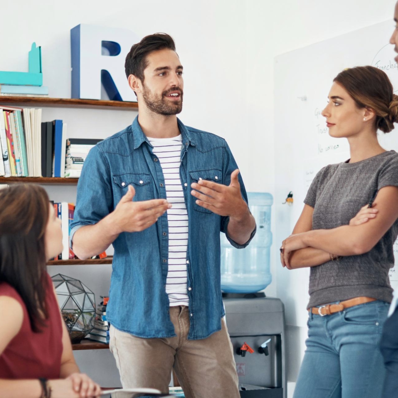 In a modern office, a white man with short dark hair and blue shirt is in discussion with three colleagues: a white woman and Black man who are stood up, and a white woman who is sat at a desk