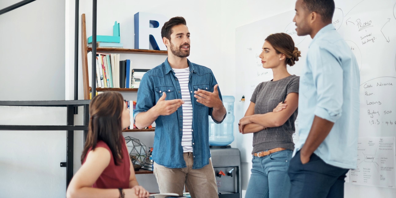 In a modern office, a white man with short dark hair and blue shirt is in discussion with three colleagues: a white woman and Black man who are stood up, and a white woman who is sat at a desk