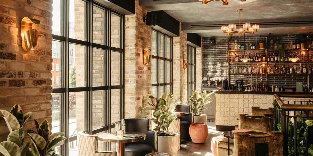 The Hoxton in Southwark - room shows tables and chairs beside a big window plants and lights and a bar in the background
