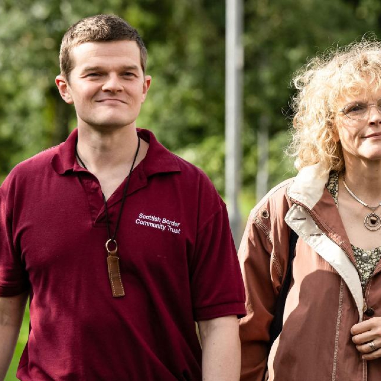 A still from the film I Swear, with actors Robert Aramayo and Maxine Peak walking along a suburban street with trees in the background.