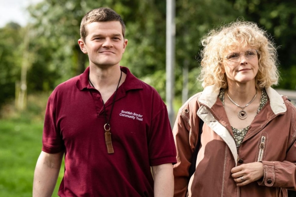 A still from the film I Swear, with actors Robert Aramayo and Maxine Peak walking along a suburban street with trees in the background.