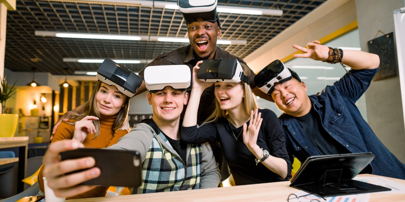Five young colleagues in a modern office smile and gesture at a phone camera while wearing virtual reality headsets on their foreheads. The colleagues are two white women, one Asian man, one Black man and one white man.