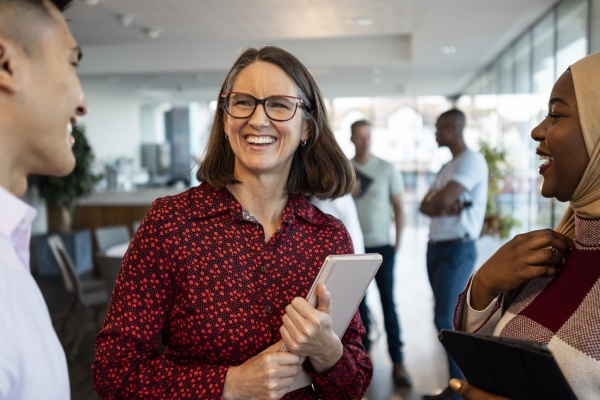 Three colleagues stood laughing together: an Asian man in pink shirt, a white woman with glasses in a red dress and a black woman wearing a beige hijab and mauve check jumper