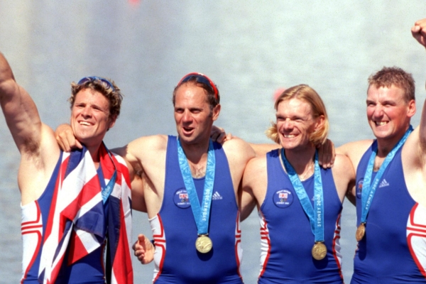 James Cracknell and team mates in a line with their arms around each other wearing blue swimsuits and displaying their gold medals with two of them punching their fists in the air
