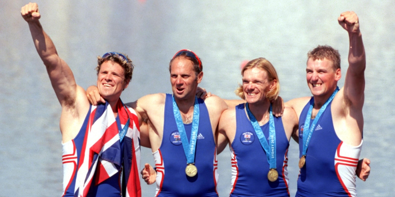 James Cracknell and team mates in a line with their arms around each other wearing blue swimsuits and displaying their gold medals with two of them punching their fists in the air