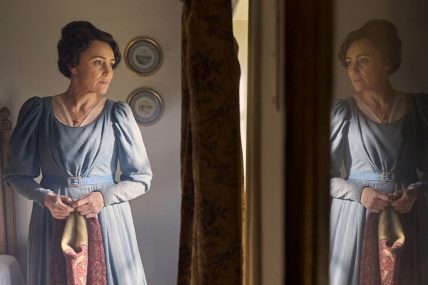 An episodic still from Miss Austen, featuring Keeley Hawes as Cassandra wearing a blue dress looking out of a bedroom window. Her reflection is on the right.