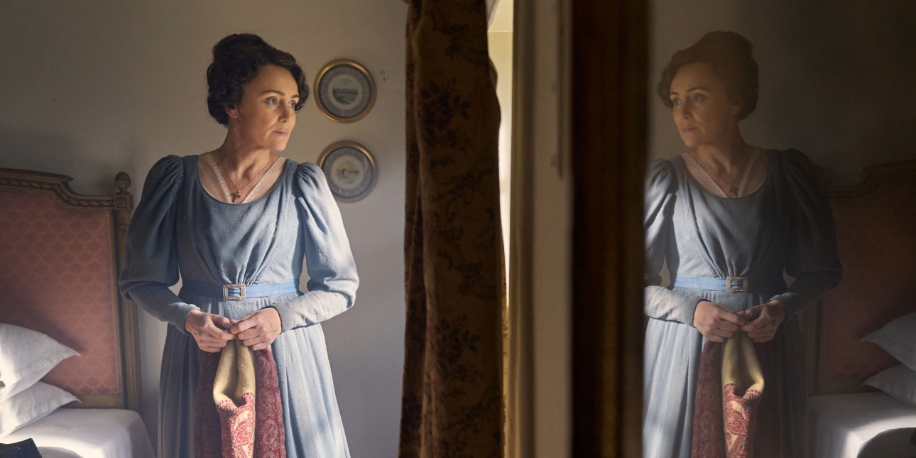 An episodic still from Miss Austen, featuring Keeley Hawes as Cassandra wearing a blue dress looking out of a bedroom window. Her reflection is on the right.