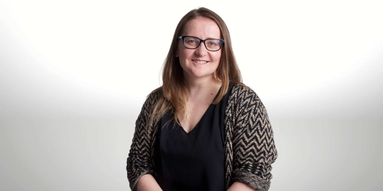 A portrait of a smiling Jule Wilson on a grey background. Jule is a white woman with long brown hair, glasses and wears a black and grey jacket over a black top