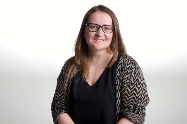 A portrait of a smiling Jule Wilson on a grey background. Jule is a white woman with long brown hair, glasses and wears a black and grey jacket over a black top