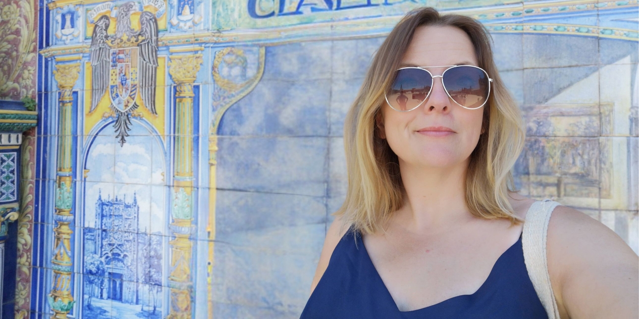 A close-up portrait of Kat Dibbits, a white woman with blonde hair and wearing glasses, against a blue and yellow tiled wall
