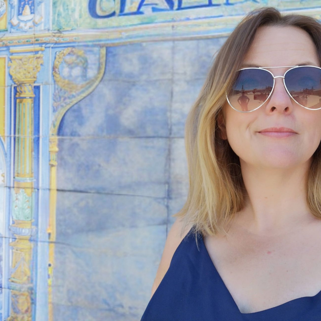 A close-up portrait of Kat Dibbits, a white woman with blonde hair and wearing glasses, against a blue and yellow tiled wall