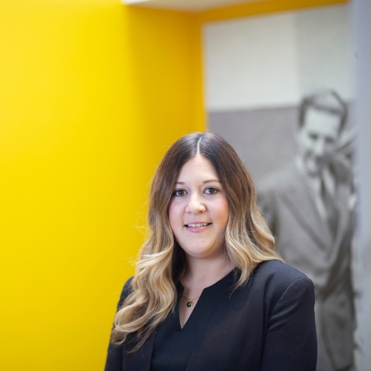 On a yellow background, a colour portrait of Laurien Hubbard. Laurien is a white woman with long, light brown hair who is wearing a black jacket. Behind her is a tall black and white photograph of a man.