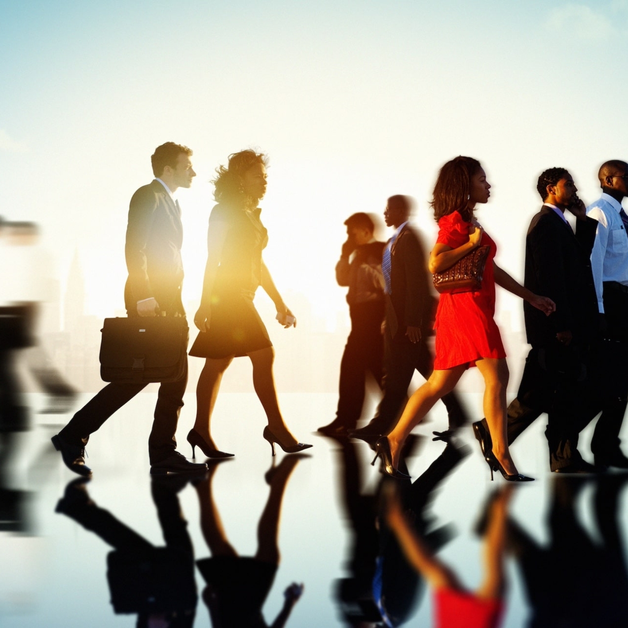 A variety of business people of different genders, ages and ethnicities walking along a reflective surface with the sun and skyscrapers behind them