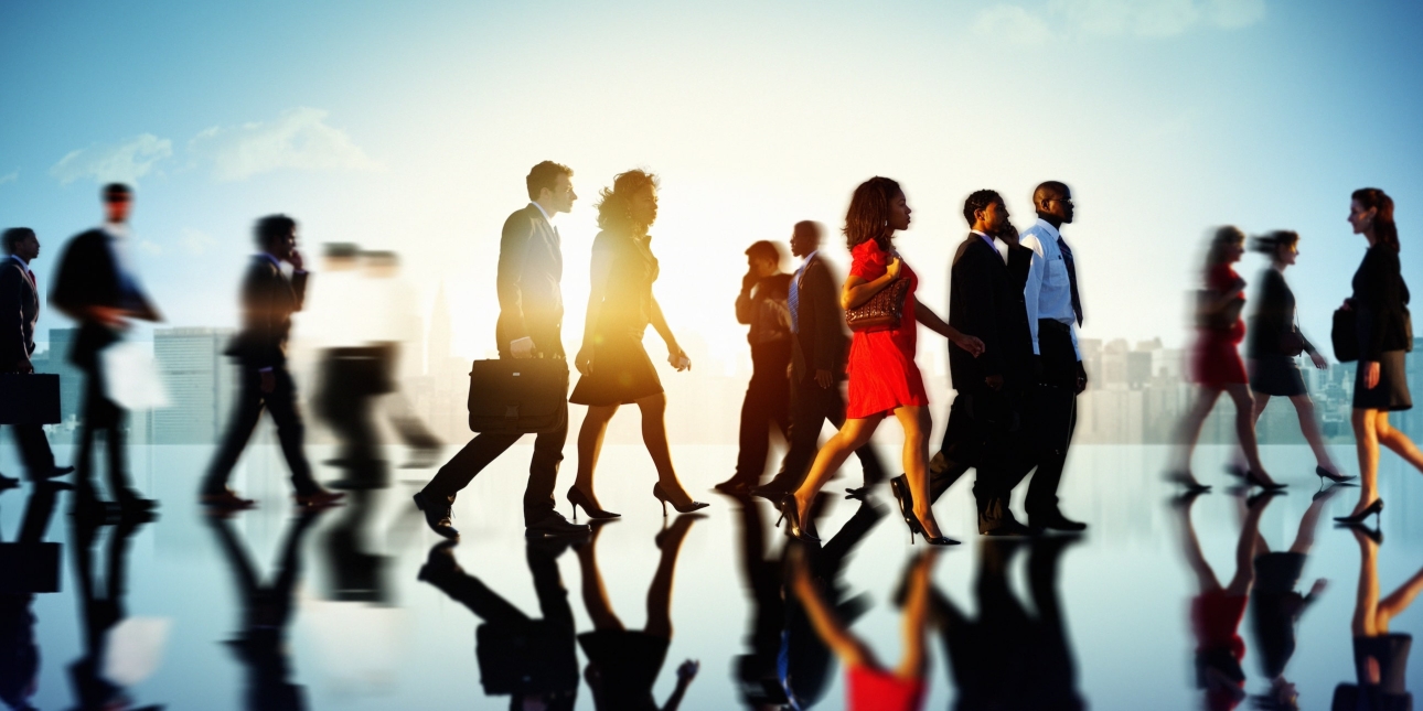 A variety of business people of different genders, ages and ethnicities walking along a reflective surface with the sun and skyscrapers behind them