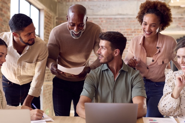 Six male and female colleagues of different ages and ethnicities gather around a laptop smiling. They are in a modern office with bare brick walls
