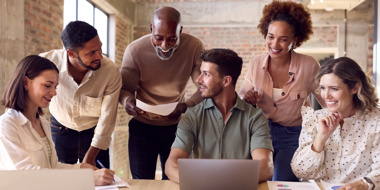 Six male and female colleagues of different ages and ethnicities gather around a laptop smiling. They are in a modern office with bare brick walls