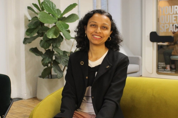 A colour portrait of Liva Emmatty sat on a mustard sofa in an office. Liva is an Asian woman with long dark hair who is wearing a black jacket over a white top and pink trousers.